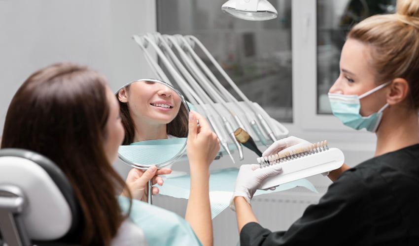 A patient sitting in a dental chair looks into a handheld mirror and smiles while a dentist wearing a mask and gloves holds a shade guide to match tooth color in a modern dental clinic.